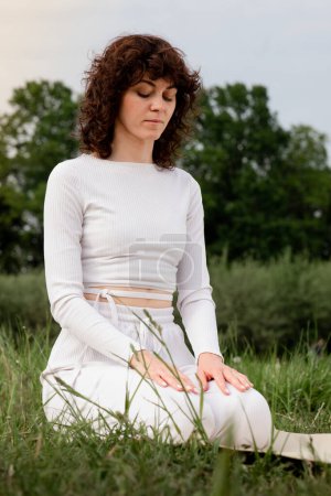Concept of calmness. Sports woman sitting on yoga mat put hut on her lap. Female focused on breathing at the park. Full concentration mindfulness. Zen posture. High quality photo
