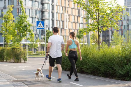 Foto de Una joven pareja atlética pasea al aire libre con un perro mascota en un cálido día de verano en la calle en un pueblo parque rodeado de coloridos edificios residenciales modernos. El concepto de caminar como un entrenamiento completo - Imagen libre de derechos
