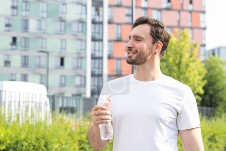 Ein positiver junger Mann joggt an einem Sommermorgen in einem modernen Wohngebiet mit bunten Gebäuden und Parklandschaft. Ein lächelnder Sportler steht zur Seite, macht eine Pause, blickt nach vorn und trinkt