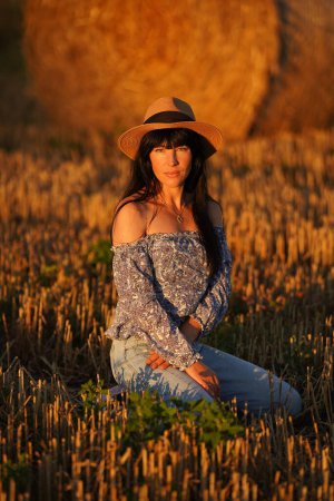 Photo for A woman with long black hair in jeans, a straw hat sits on her knees in a field in front of a haystack, her blouse lowered and her shoulders bare, she looks into the camera lens. Brunette in the field - Royalty Free Image