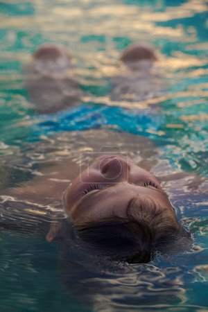 Téléchargez les photos : Le garçon est allongé dans de l'eau turquoise sur le dos les yeux fermés. Garçon relaxant à la piscine de l'hôtel en été. - en image libre de droit