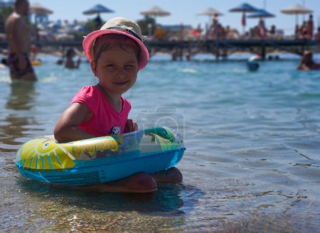 Foto de Una niña con un sombrero de paja se sienta en el agua junto al mar y entrecierra los ojos al sol. Fondo borroso. Pequeña linda chica se sienta en la playa. - Imagen libre de derechos