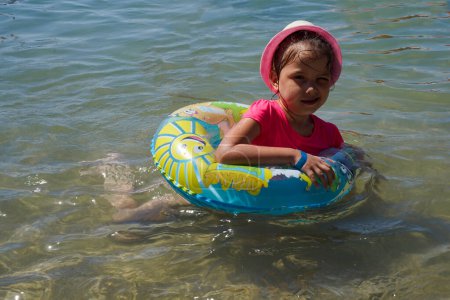 Foto de Una niña con un sombrero de paja se sienta en el agua en el mar. Pequeña linda chica se sienta en la playa. - Imagen libre de derechos