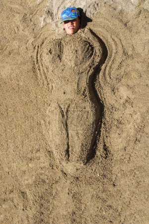 Téléchargez les photos : La tête du garçon sort du sable, le corps est enterré dans le sable, les plaisirs de l'été sur la plage. Mignon garçon enterré dans le sable pour le plaisir - en image libre de droit