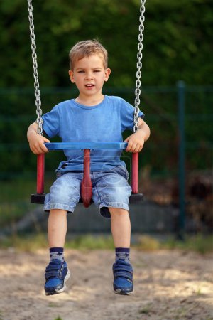 Téléchargez les photos : Un petit garçon est assis sur une balançoire. Enfant mignon jouant sur swing dans la cour arrière à la campagne - en image libre de droit