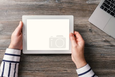 Photo for A middle-aged man holds a tablet in his hands. Against the background of a beautiful wooden table with a laptop. Template for the designer. - Royalty Free Image