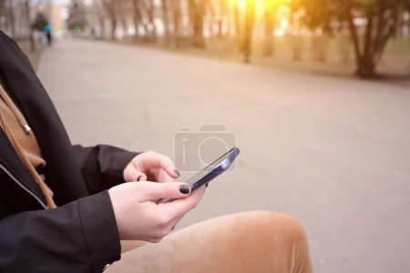 A girl sits on a bench in a city park with a smartphone in her hands. Empty paths, loneliness concept.