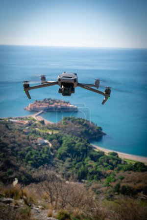 Focused close-up of a drone hovering with spinning blades, with a blurry coastal landscape and island in the background. 