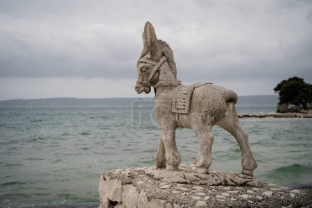 Eine verwitterte Steinskulptur eines Esels steht auf einem Küstenpier mit wolkenverhangener Meereskulisse. 