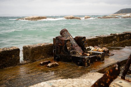 An old, corroded winch stands on a wet dock, with choppy sea waves and rocky outcrops in the background. 