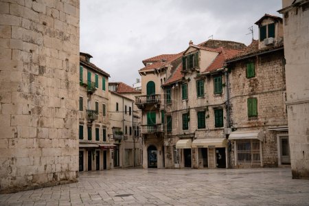 A serene, empty town square in an old European city, surrounded by weathered stone buildings with green shutters. 
