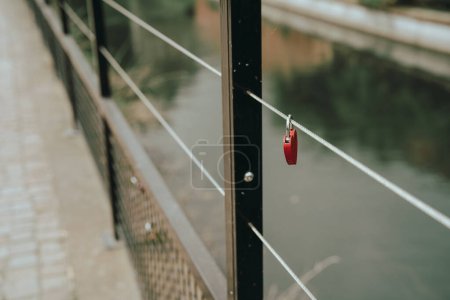 Un cadenas rouge unique, symbolisant souvent l'amour durable, est attaché à la balustrade métallique d'un pont au-dessus des eaux calmes. 
