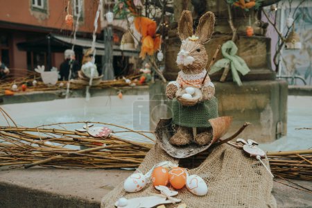A straw Easter bunny holding eggs sits atop a burlap cloth, part of an elaborate festive display in a town square setting. 