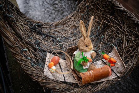 An adorable Easter bunny figurine adorned with a festive outfit sits in a wicker basket alongside colorful eggs and carrots. 