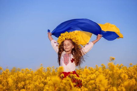 Ukrainian child girl in embroidered shirt and yellow wreath in field of yellow flowers against blue sky. Ukraine s blue-yellow flag flying in wind in hands of little Ukrainian girl. Pray for Ukraine.