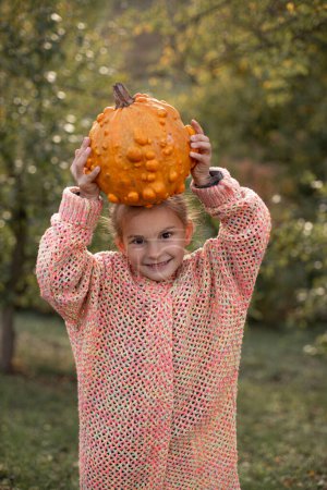 Photo for Happy smilling child girl in a knitted sweater is holding in hands ugly orange pumpkin. Deformed orange pumpkin with a damaged, ugly skin. Thanksgiving, harvest, halloween concept. - Royalty Free Image