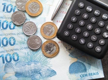 Concepto financiero billetes y monedas de cien reales con calculadora y pluma de dinero brasileño.