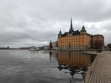 A scenic view of Stockholm's waterfront featuring historic buildings reflected in the calm waters under an overcast sky