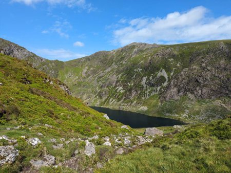 Landschaftliche Berglandschaft mit einem ruhigen See in Snowdonia, Wales. Felsiges Gelände und üppiges Grün unter strahlend blauem Himmel. Perfekt für Outdoor-Abenteuer, Natur-Inspiration und friedlichen Rückzug.