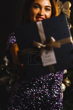 Téléchargez les photos : Une jeune belle femme dans une robe de soirée élégante et brillante bleue se tient près du sapin de Noël et ouvre des cadeaux la veille du Nouvel An. Intérieur avec décorations de Noël. - en image libre de droit
