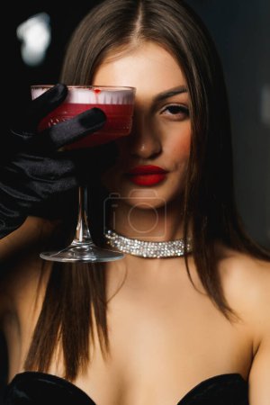 Téléchargez les photos : Femme sexy dans une robe noire avec des gants et des lèvres rouges tient un cocktail.Nightlife concept - en image libre de droit