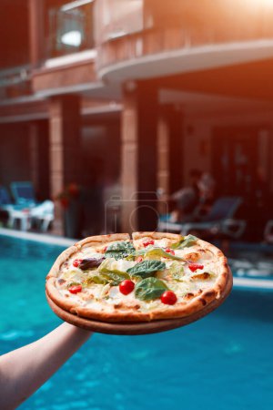 Foto de Una mano sostiene una pizza sobre el fondo de una piscina. El concepto de pizza y vacaciones en el hotel - Imagen libre de derechos