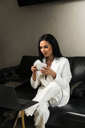 Eine Geschäftsfrau im weißen Anzug sitzt im Büro am Laptop und trinkt Kaffee. Büroarbeitskonzept. Fernbedienung auf einem PC