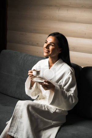 Junge Frau im weißen Bademantel trinkt Espresso in einem Hotelzimmer aus Holz. Hotelurlaubskonzept. Kaffee am Morgen
