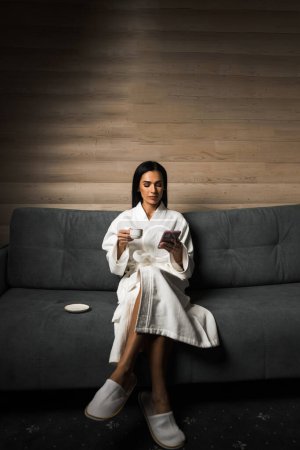 Junge Frau im weißen Bademantel trinkt Espresso in einem Hotelzimmer aus Holz. Hotelurlaubskonzept. Kaffee am Morgen