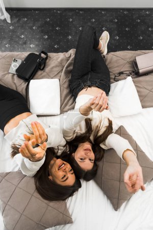 Deux jeunes femmes partagent le bonheur et prennent un selfie au lit dans la chambre. Concept vacances hôtel