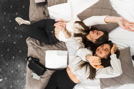 Two young women share happiness and take a selfie in bed in the room. Hotel holiday concept