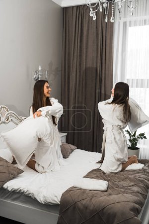 Téléchargez les photos : Deux jeunes femmes en peignoir riant et frappant des oreillers au lit. Concept vacances hôtel - en image libre de droit