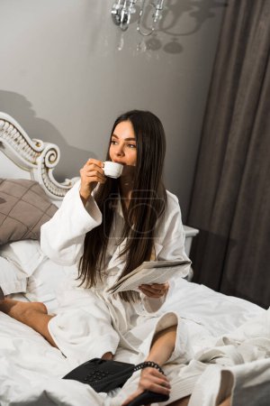 Junge Frauen in Bademänteln frühstücken im Bett, trinken Kaffee und albern herum. Frühstück Käsekuchen und Obst mit Kaffee. Hotelurlaubskonzept