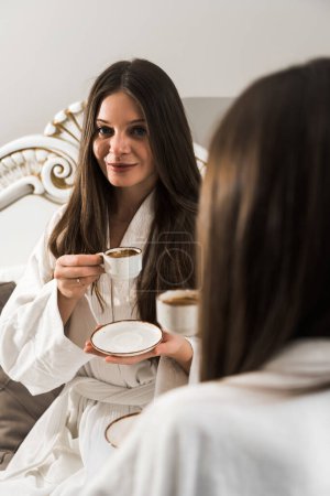 Junge Frauen in Bademänteln frühstücken im Bett, trinken Kaffee und albern herum. Frühstück Käsekuchen und Obst mit Kaffee. Hotelurlaubskonzept