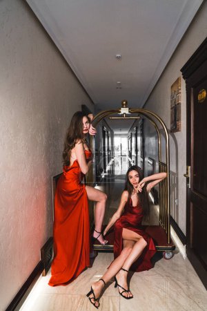 Téléchargez les photos : Deux femmes sexy en robes rouges montent sur un chariot à bagages doré dans un hôtel. Concept de tir de mode - en image libre de droit