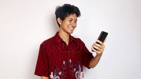 Schöner junger asiatischer Mann in Batik zeigt ein glückliches Lächeln auf seinem Smartphone auf einem isolierten weißen Hintergrund