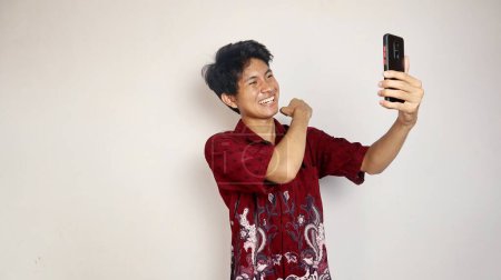 happy young handsome asian man in batik shirt posing for selfie on smartphone on isolated white background