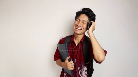 glücklicher junger gutaussehender asiatischer Mann im Batikhemd, der eine Tasche in der Hand hält und mit einem Buch posiert, während er mit einem Smartphone auf einem isolierten weißen Hintergrund telefoniert