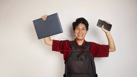 Handsome young Asian man in batik shirt carrying a bag, holding a book and smartphone, expressing joy, happiness, enthusiasm, victory on an isolated white background