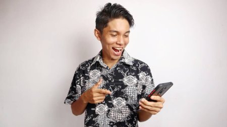 Happy young handsome Asian man wearing batik shirt posing pointing and holding smartphone on isolated white background
