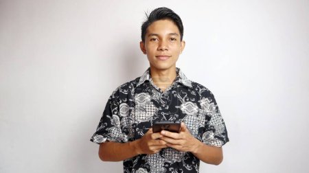 Happy young handsome Asian man wearing batik shirt posing holding smartphone on isolated white background