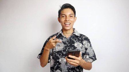 Glücklicher junger gutaussehender asiatischer Mann in Batikhemd, der mit einem Smartphone auf isoliertem weißem Hintergrund posiert