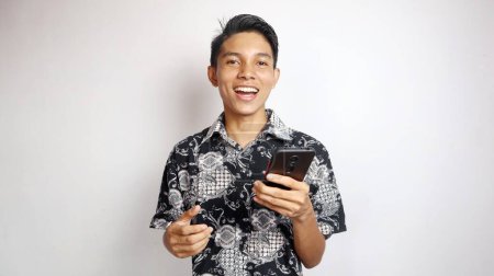 Glücklicher junger hübscher asiatischer Mann in Batikhemd posiert mit Smartphone auf isoliertem weißem Hintergrund