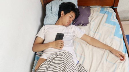 A young Asian man in a white shirt is asleep on the bed and still holding a smartphone