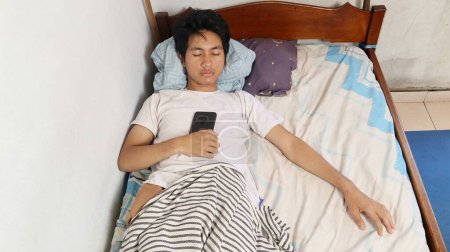 A young Asian man in a white shirt is asleep on the bed and still holding a smartphone