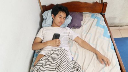 Un joven asiático con una camisa blanca está dormido en la cama y todavía sostiene un teléfono inteligente