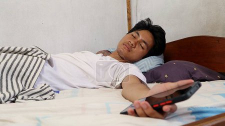 Ein junger Asiate in weißem Hemd schläft auf dem Bett und hält immer noch ein Smartphone in der Hand