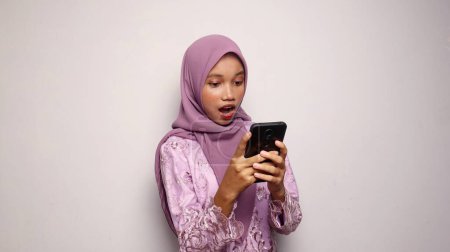 schockiert schöne indonesische Teenager-Mädchen trägt einen Kebaya und Hijab hält und schaut auf ein Smartphone auf einem isolierten weißen Hintergrund