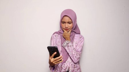 Schöne indonesische Teenager-Mädchen tragen Kebaya und Hijab halten und Blick auf Smartphone auf isoliertem weißen Hintergrund