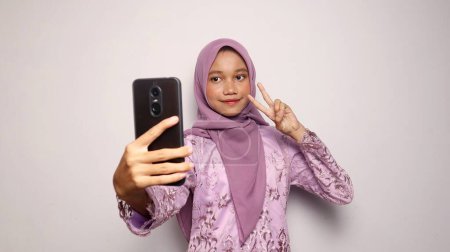 Indonesian teenage girls wearing kebaya and hijab pose holding smartphone selfies on an isolated white background